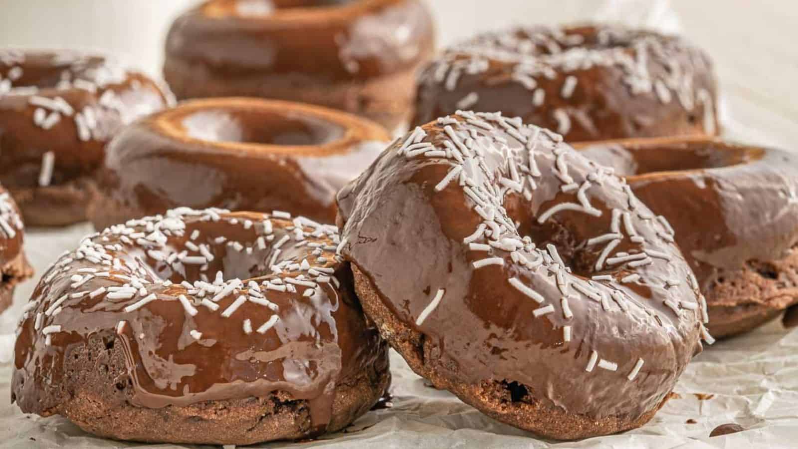 A group of chocolate-glazed donuts topped with white sprinkles are arranged on a sheet of parchment paper.