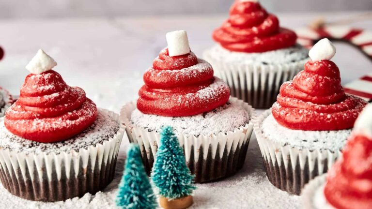 Chocolate cupcakes with red swirled frosting, dusted with powdered sugar, each topped with a small marshmallow, placed beside miniature decorative trees.