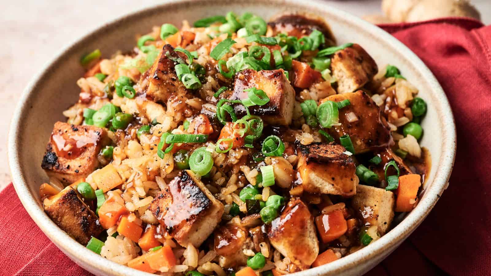 A bowl of fried rice with grilled tofu, diced carrots, peas, chopped green onions, and a drizzle of dark sauce, served on a red cloth.