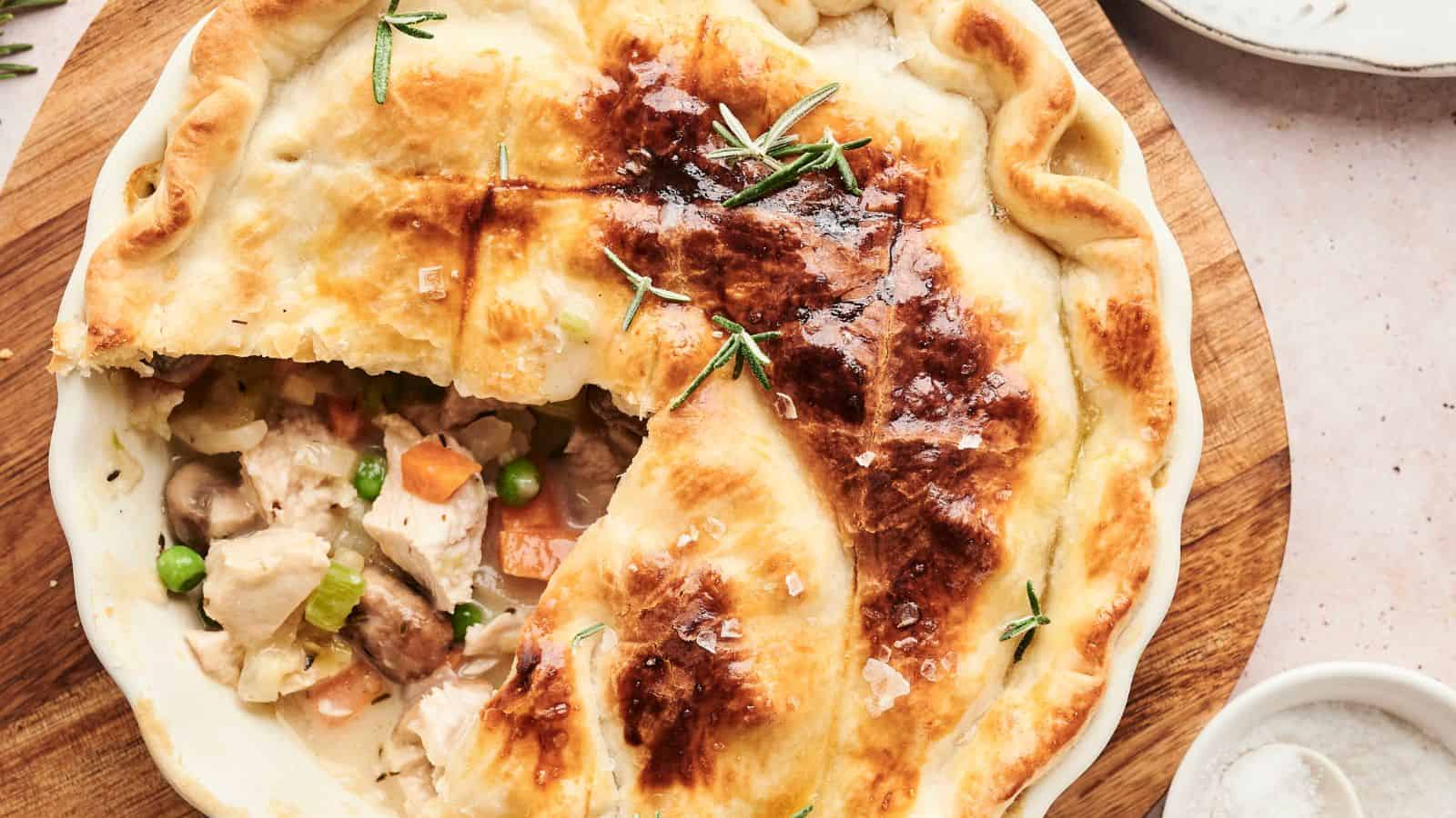 A steaming chicken pot pie with a golden-brown crust, partially cut open to reveal chunks of chicken, peas, carrots, and mushrooms. Sprigs of rosemary garnish the top.