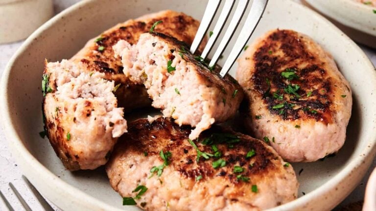 Four browned sausage patties garnished with chopped herbs on a plate, with a fork breaking one open to show the inside.