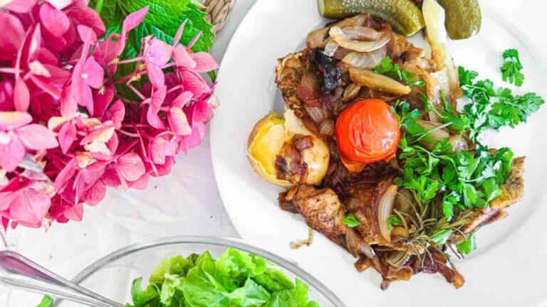 A plate with roasted vegetables and pork fillet, including tomato, onion, and potatoes, garnished with fresh parsley, with pickles on the side. Pink flowers and a bowl of lettuce are nearby.