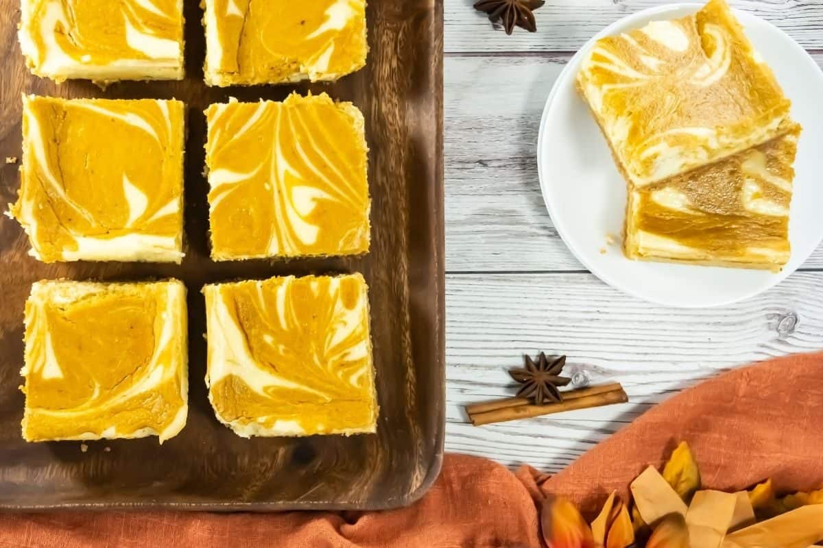Six square pumpkin cheesecake bars are arranged on a wooden tray, displaying marbled orange and white patterns on top.