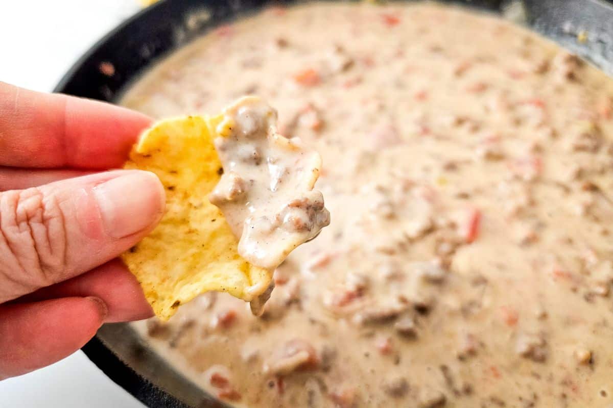 A hand holding a tortilla chip with creamy Queso Blanco Rotel Dip, over a skillet filled with the same dip.