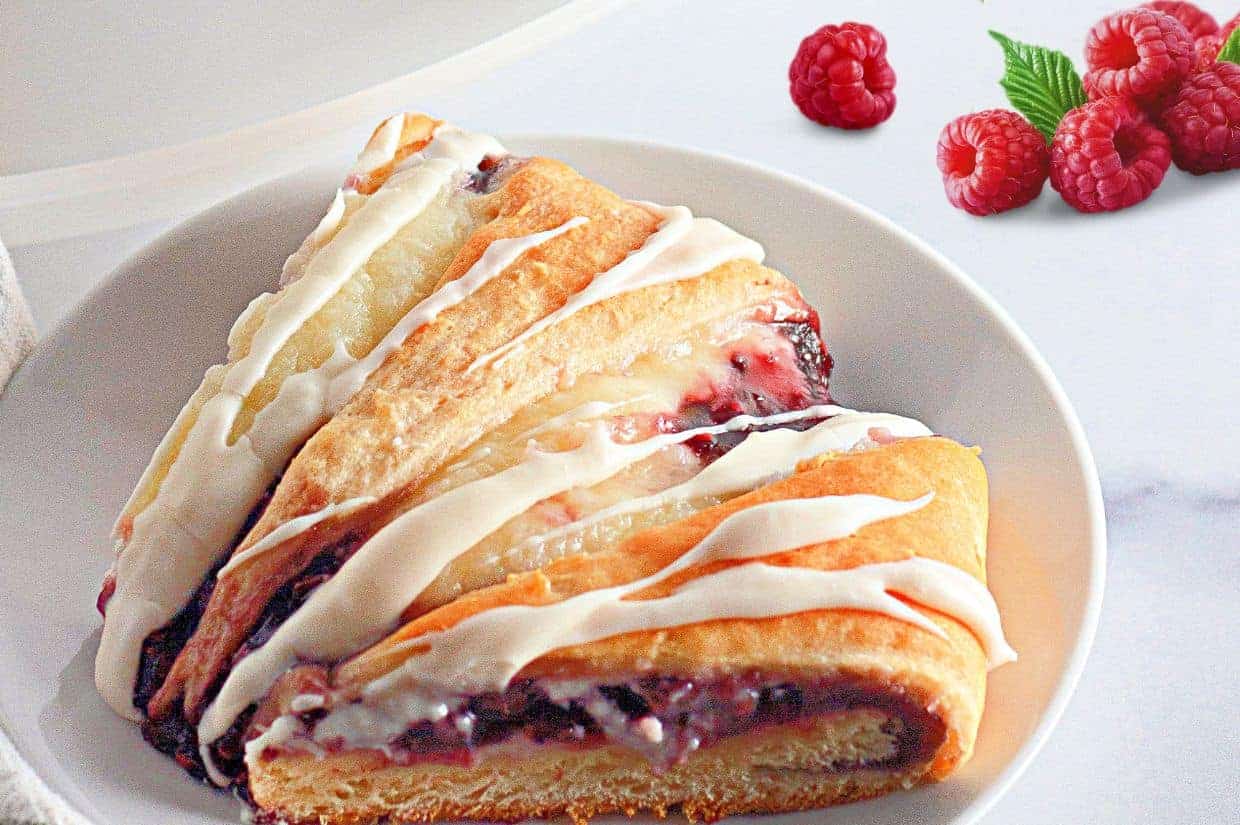 A slice of raspberry crescent ring with white icing on a white plate, with fresh raspberries and a green leaf in the background.