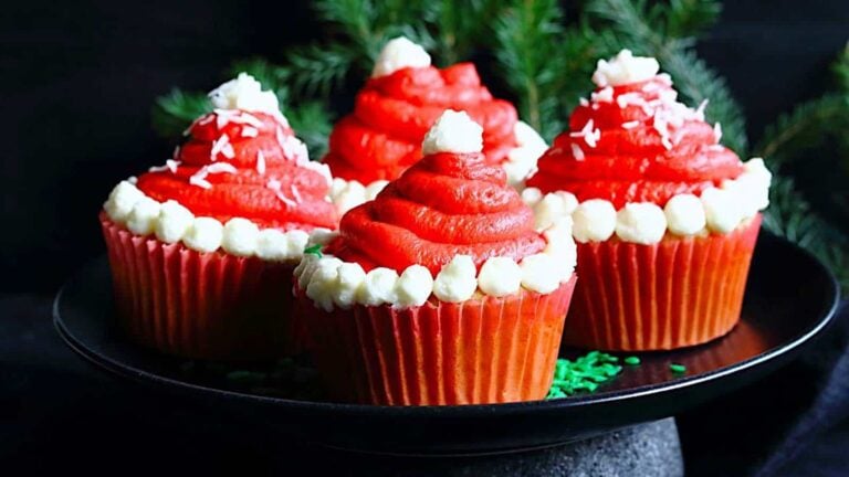 Four cupcakes decorated with red and white frosting resembling Santa hats are arranged on a black plate, with green pine branches in the background.