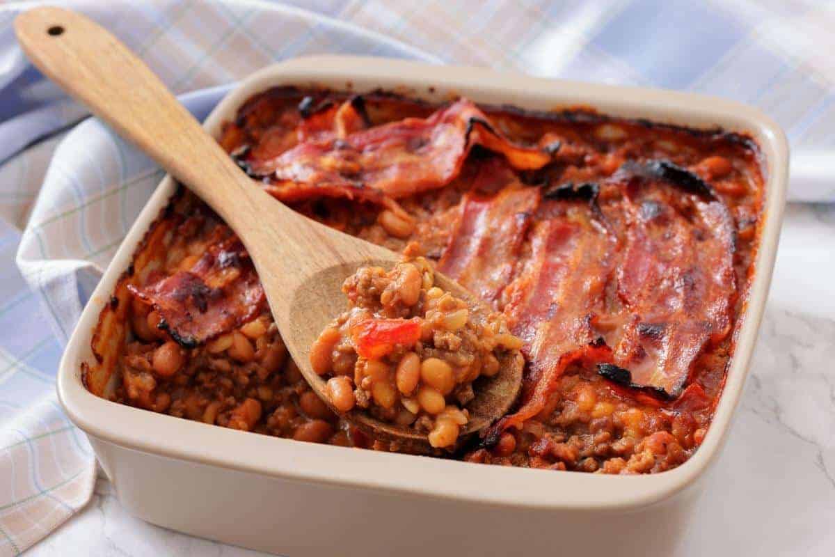 A square baking dish filled with Southern Ground Beef & Baked Bean Casserole and topped with strips of cooked bacon; a wooden spoon holds a serving.