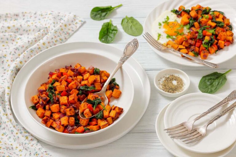 A white table with bowls and plates of sweet potato hash with spinach and red onion, surrounded by fresh spinach leaves, a napkin, pepper, and forks.