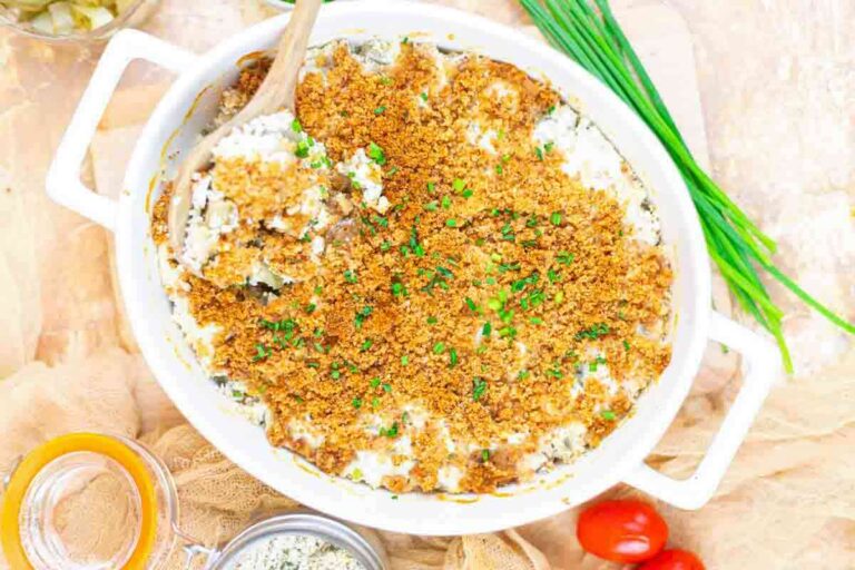 A white casserole dish filled with a baked creamy dip topped with golden breadcrumbs and chopped chives, with a wooden spoon placed inside.