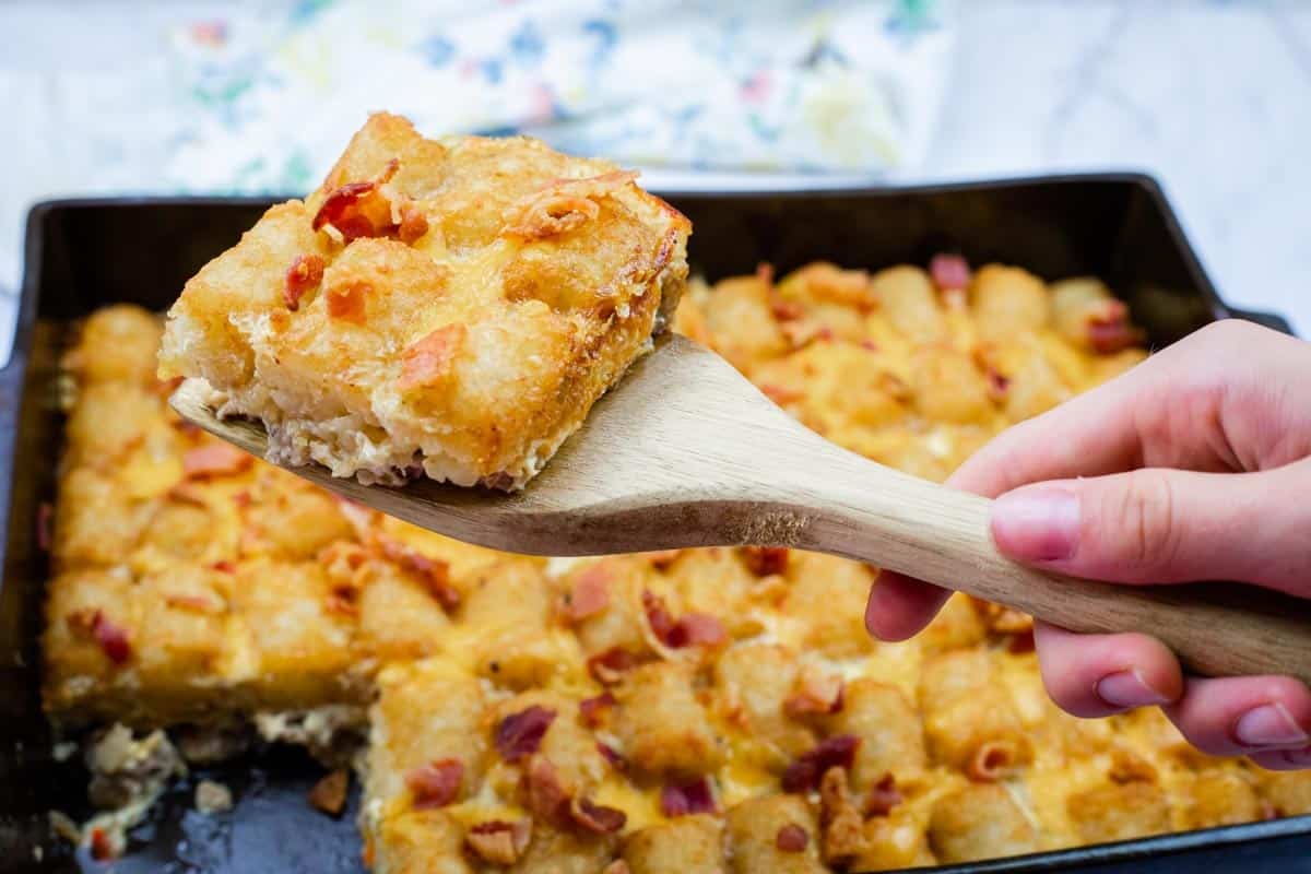 A wooden spatula holds up a square piece of Tater Tot Breakfast Casserole topped with crispy bits.