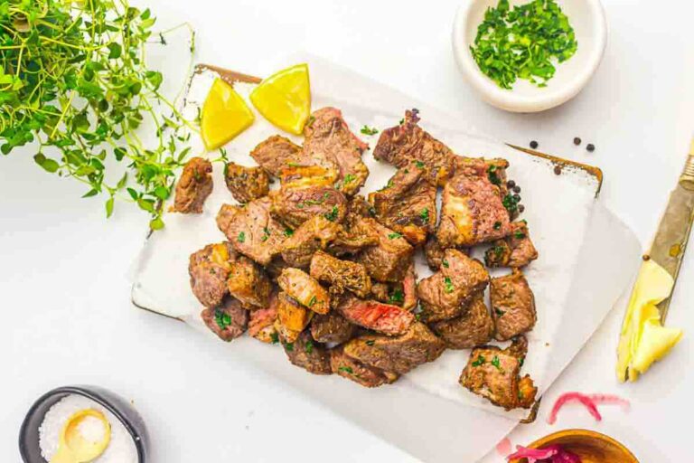 Air Fryer beef steak bites garnished with herbs, served with lemon wedges on a white tray, surrounded by seasonings and fresh herbs.