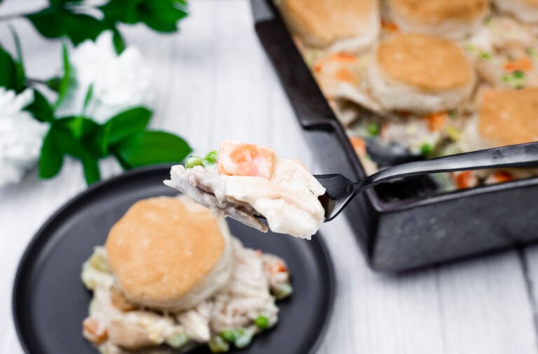 A fork holds a bite of chicken pot pie with vegetables and biscuit topping, with a plate and baking dish of the same meal in the background.