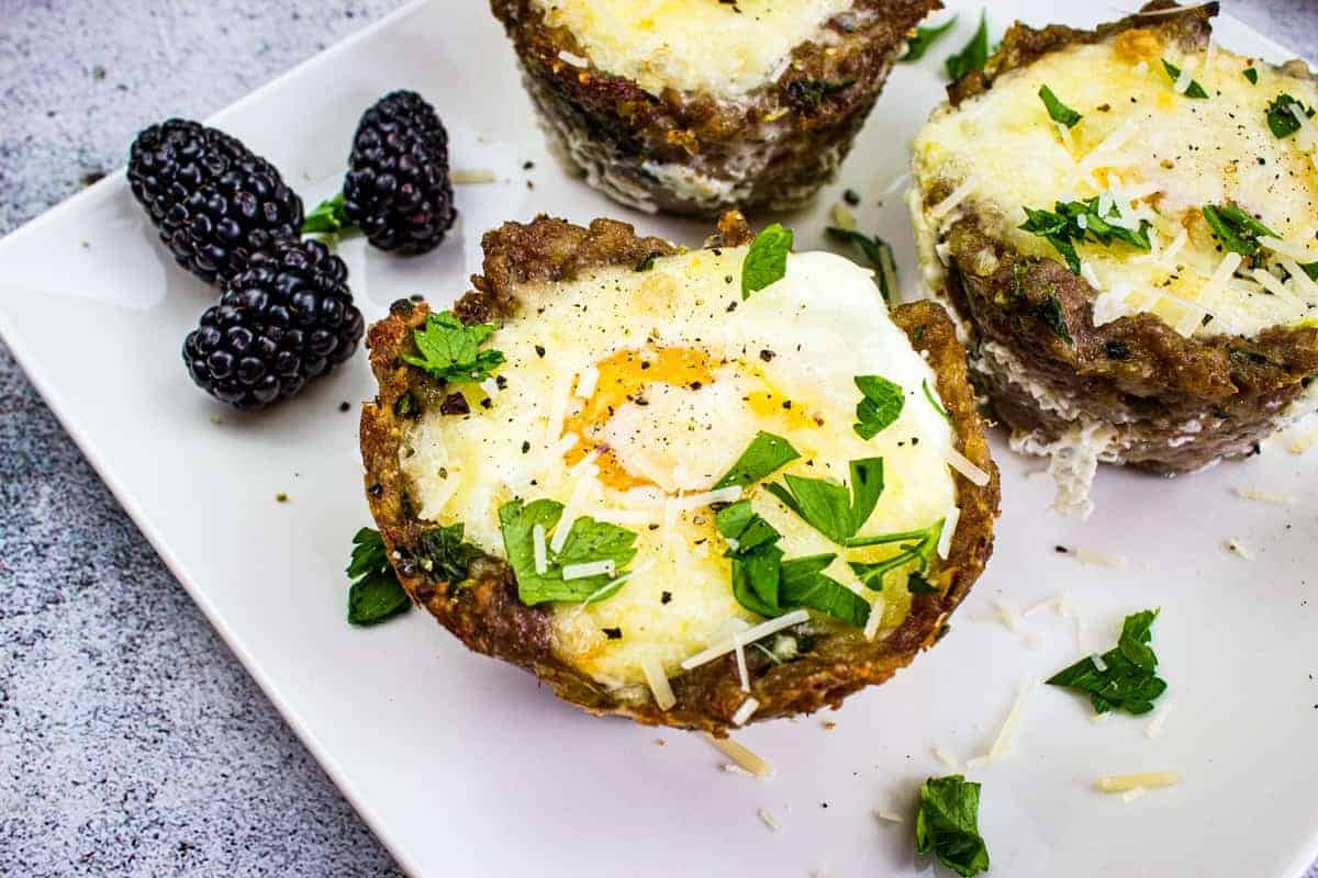 Three baked egg and meat cups topped with herbs and cheese are on a white plate, accompanied by blackberries.