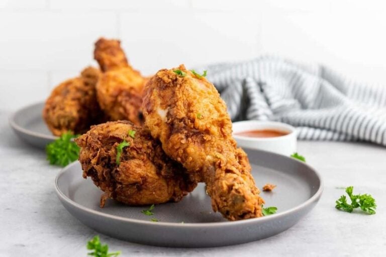 Three pieces of fried chicken drumsticks on a gray plate, garnished with parsley, with a cup of dipping sauce and a striped cloth in the background.