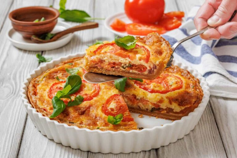 A hand lifts a slice of tomato and cheese quiche from a ceramic dish, with fresh tomatoes and herbs on a wooden table in the background.