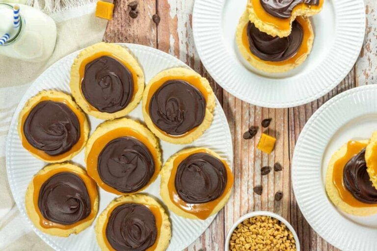 A plate of cookies topped with caramel and chocolate icing, next to a plate with two stacked cookies, a bowl of toffee bits, and a small bottle of milk on a wooden table.