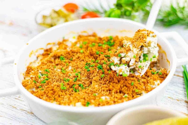 A white casserole dish filled with a baked creamy mixture topped with golden brown breadcrumbs and chopped chives, with a serving spoon lifting a portion.