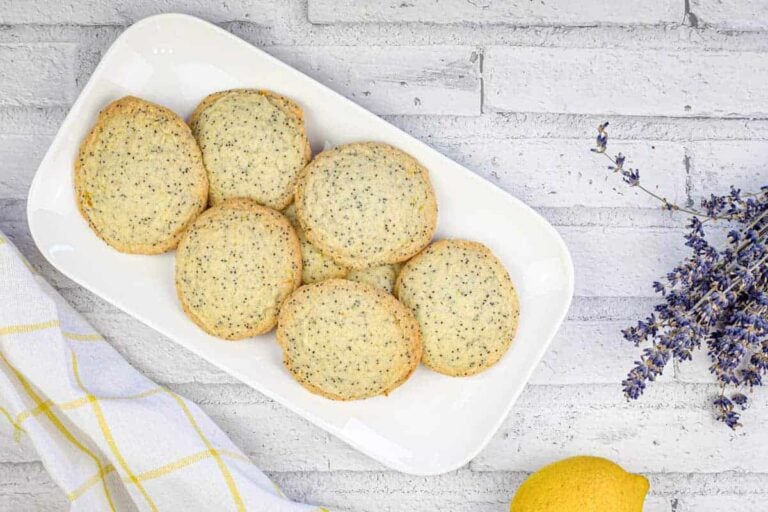 A white rectangular plate with seven poppy seed cookies sits on a light gray brick surface, next to a bunch of lavender, a lemon, and a white towel with yellow stripes.