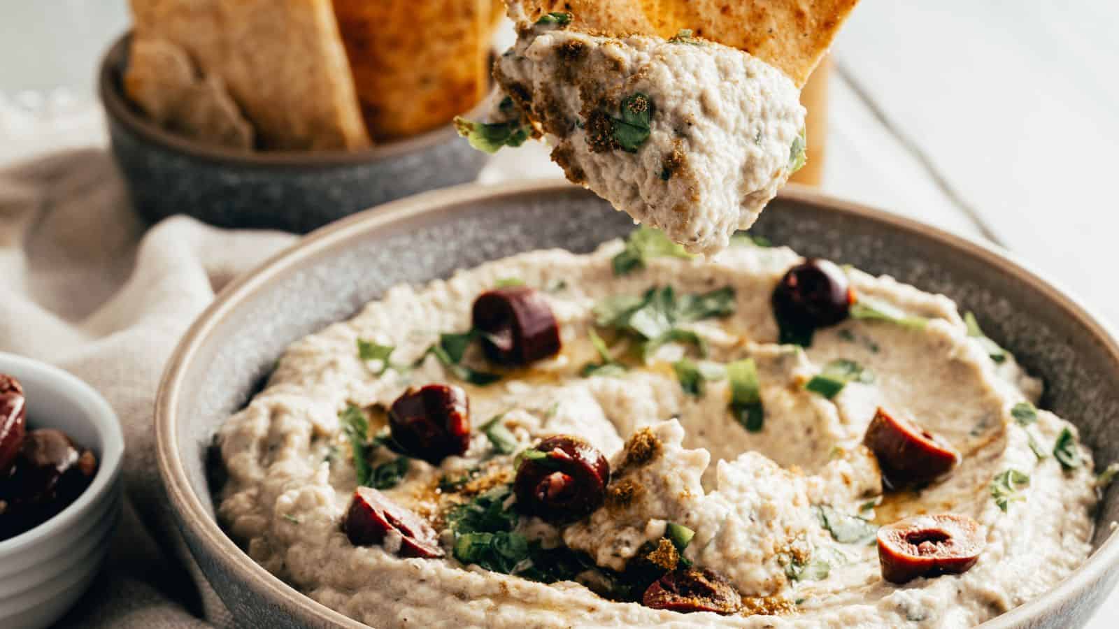 A bowl of baba ganoush topped with chopped herbs and olives, with a pita chip being dipped into the spread.