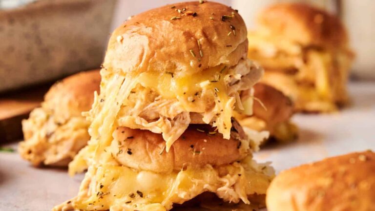 Close-up of stacked sliders filled with shredded chicken and melted cheese, with herbs sprinkled on top and more sliders blurred in the background.