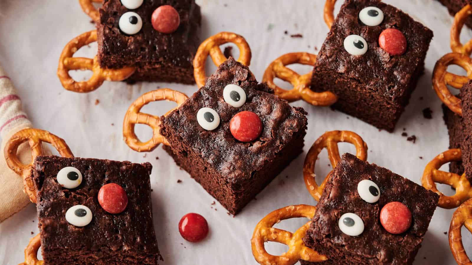 Several brownies decorated with candy eyes, red candy noses, and pretzel antlers are arranged on a white surface to resemble reindeer faces.