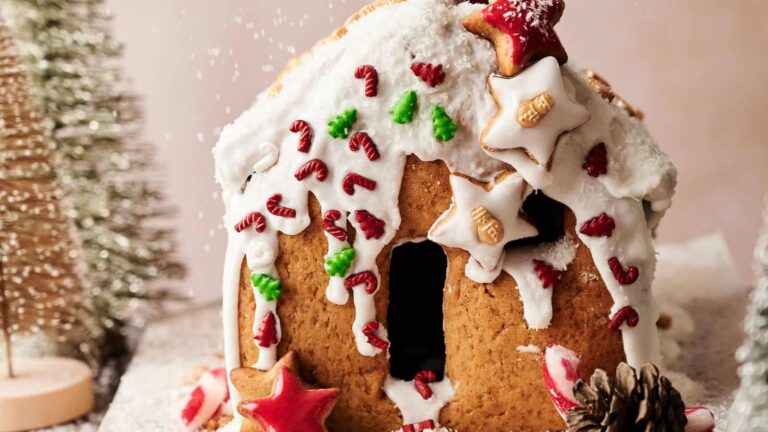 A gingerbread house decorated with white icing, star-shaped cookies, and red and green candy, with artificial snow sprinkled on top.