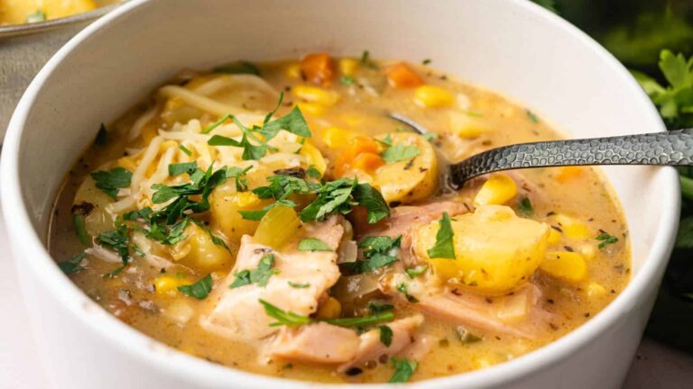 A close-up of a bowl of chicken and vegetable chowder with potatoes, corn, carrots, and chopped parsley, with a spoon inside the bowl.