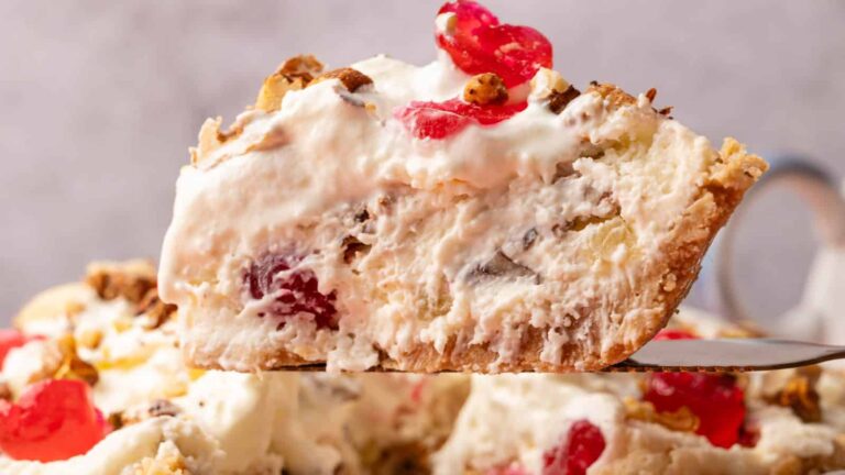A slice of creamy pie with visible cherries, nuts, and a flaky crust is held up against a blurred background.