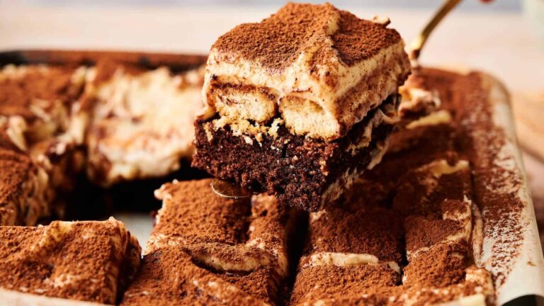 A serving of layered tiramisu with cocoa powder on top is lifted from a tray of sliced tiramisu.