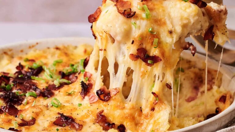 A serving of cheesy baked casserole is being lifted from a dish, showing melted cheese, crispy bacon, and chopped green onions on top.