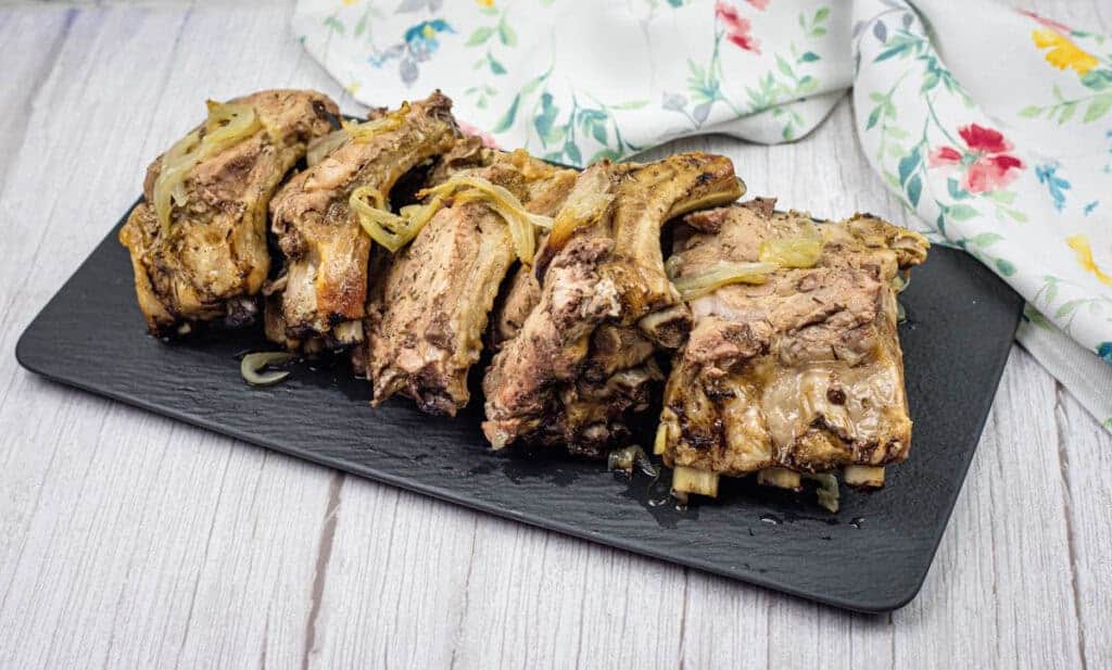 Oven-baked ribs with onion and garlic on top served on a white rectangular plate.