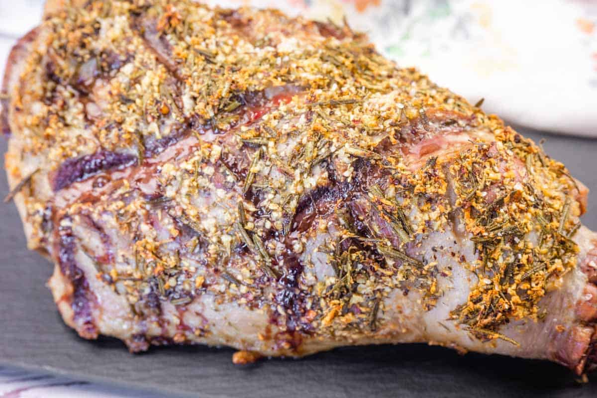 Close-up of a cooked, roasted leg of lamb coated with a crust of herbs and spices, resting on a dark surface.