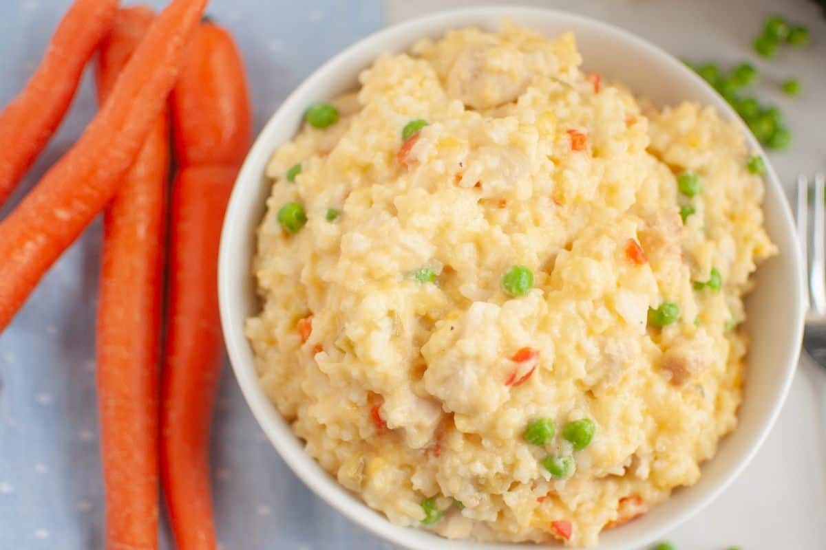 A bowl of Slow Cooker Chicken and Rice is next to whole carrots on a light blue cloth.