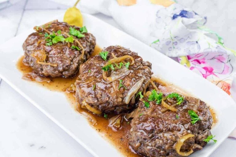 Three servings of Salisbury steak topped with onions and brown gravy are arranged on a white rectangular plate, garnished with chopped parsley.