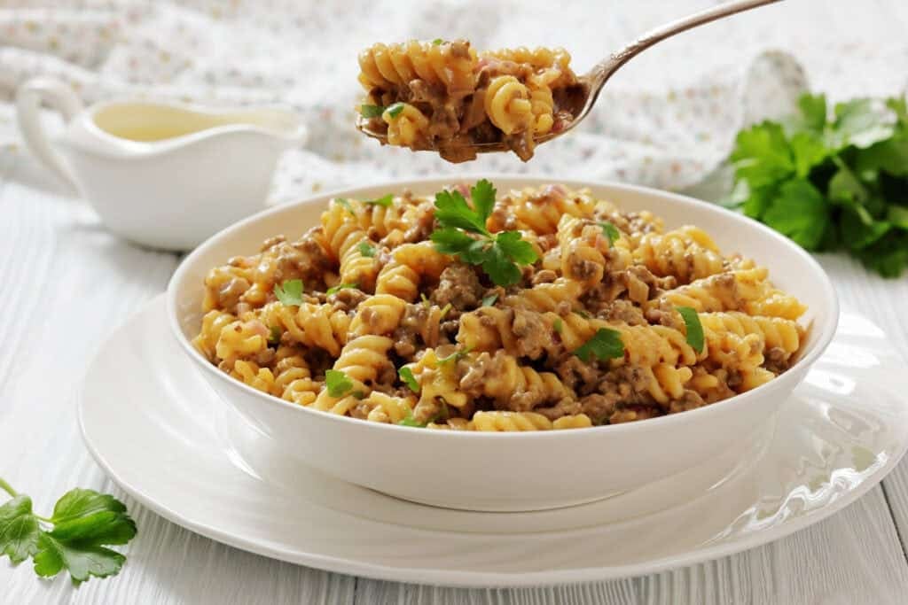 A white bowl of Cheeseburger Hamburger Helper, garnished with parsley, sits on a white plate, with a fork holding a bite above the bowl.
