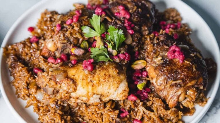 A plate of seasoned rice topped with pieces of cooked chicken, garnished with pomegranate seeds, parsley, and pistachios.