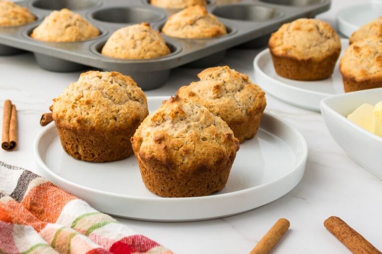 Three baked muffins are on a white plate, with more muffins in the background in a muffin tin. A patterned cloth and cinnamon sticks are nearby.