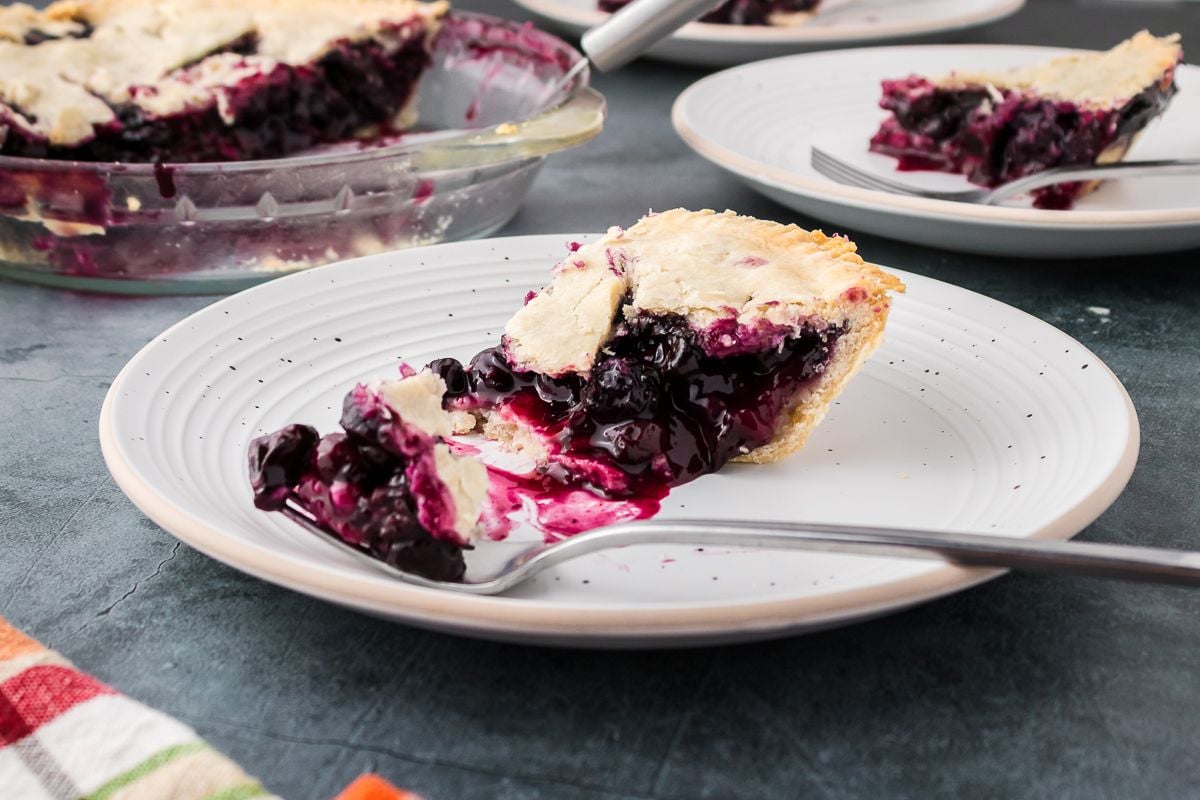 A slice of blueberry pie with a golden crust and visible berry filling sits on a white plate; additional pie slices are in the background.