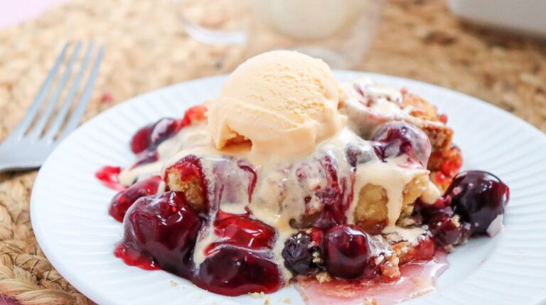 A serving of cherry cobbler topped with a scoop of vanilla ice cream is on a white plate with a fork beside it.