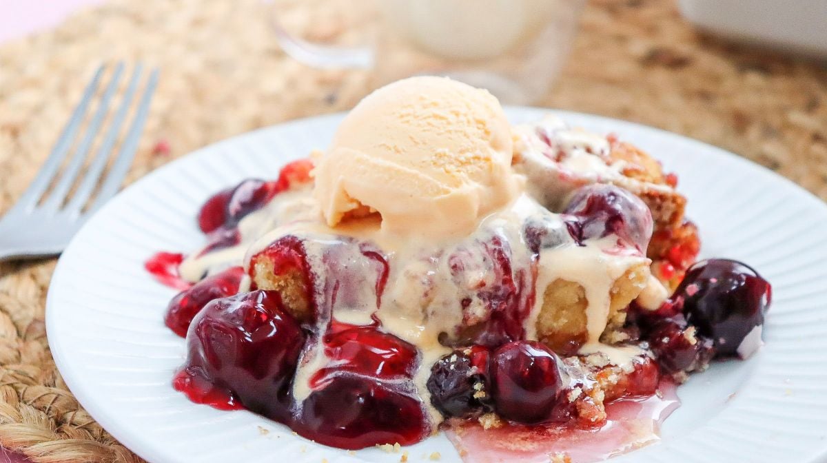 A serving of cherry cobbler topped with a scoop of vanilla ice cream is on a white plate with a fork beside it.