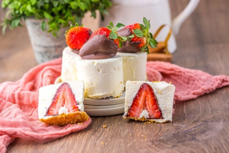 A small cheesecake topped with chocolate-covered strawberries, with two slices showing a large strawberry inside, placed on a wooden surface with a pink cloth.