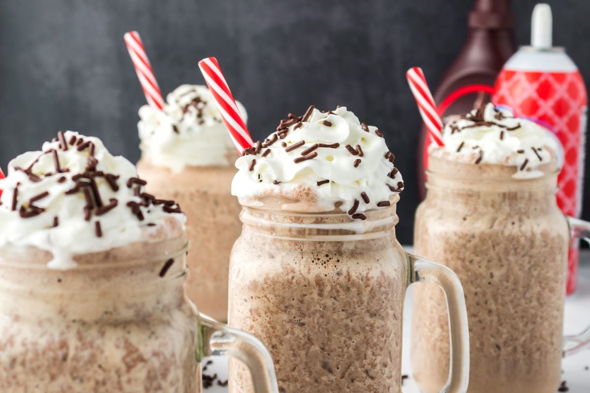 Four chocolate milkshakes in mason jars topped with whipped cream, chocolate sprinkles, and red striped straws, with chocolate syrup and whipped cream cans in the background.