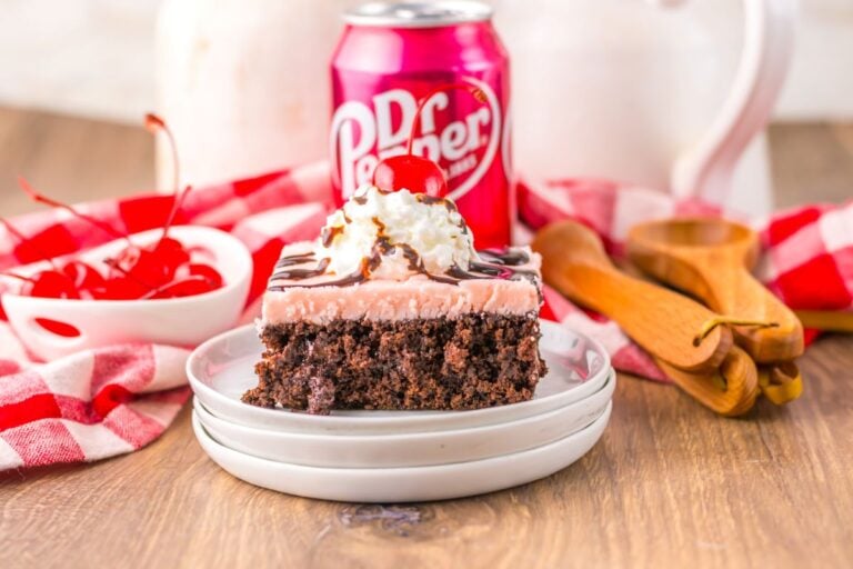 A slice of chocolate cake with pink frosting, whipped cream, chocolate drizzle, and a cherry on top sits on a plate beside a can of Dr Pepper and a bowl of cherries.