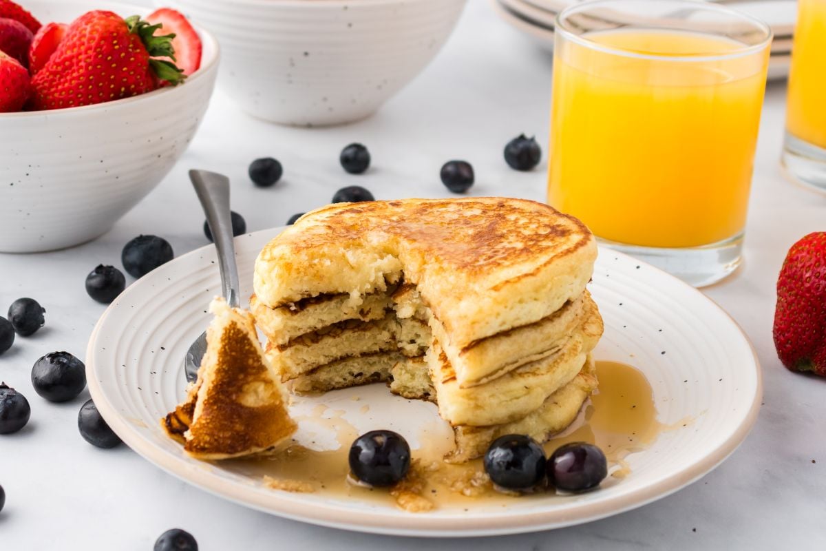 A stack of pancakes with syrup and blueberries on a white plate, a fork taking a piece, with a glass of orange juice and a bowl of strawberries in the background.
