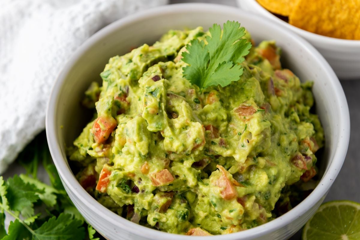 A bowl of guacamole garnished with a cilantro leaf, surrounded by lime slices and tortilla chips on the side.