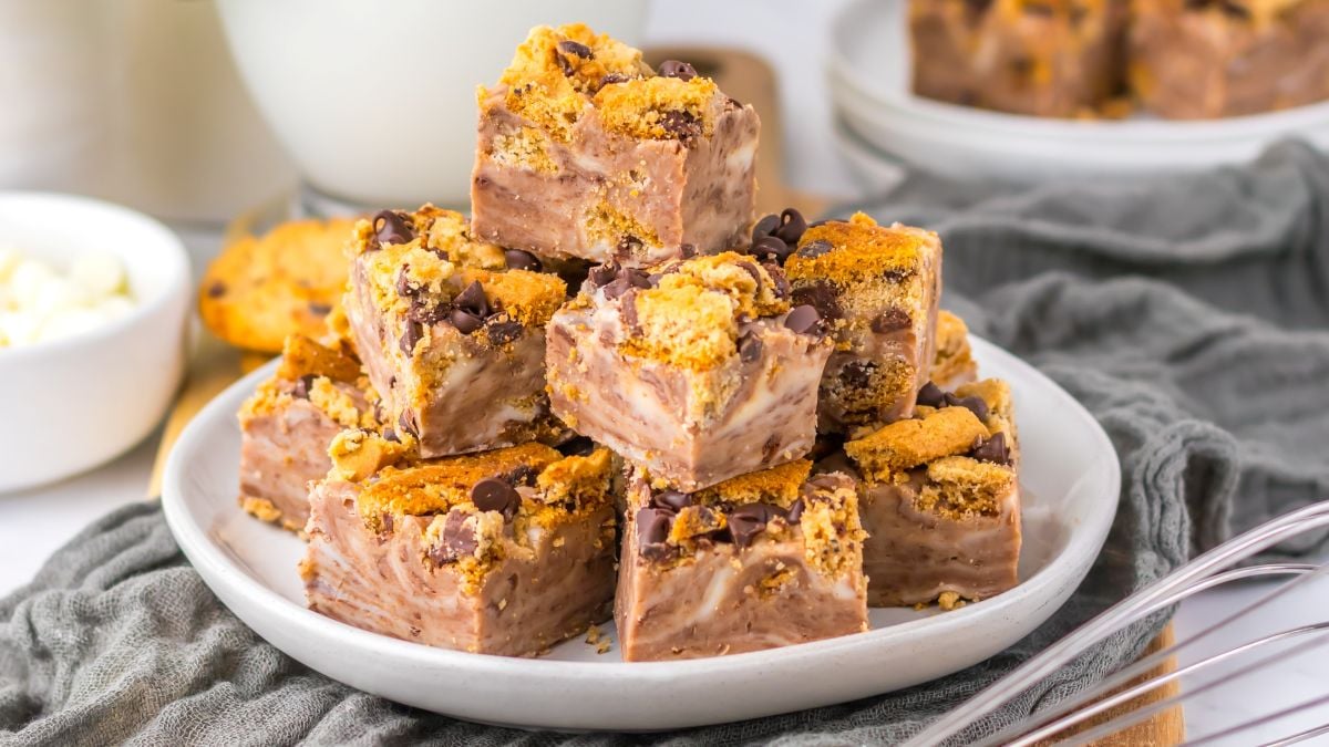 A plate stacked with fudge squares topped with crumbled cookies and chocolate chips, set on a gray cloth with a blurred background including a glass of milk and a bowl of white candies.