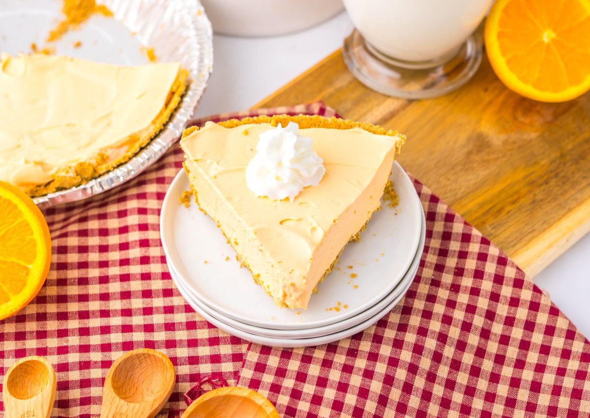 A slice of creamy pie with whipped cream on top sits on stacked plates beside a whole pie, wooden spoons, orange slices, and a glass of milk on a red checkered cloth.