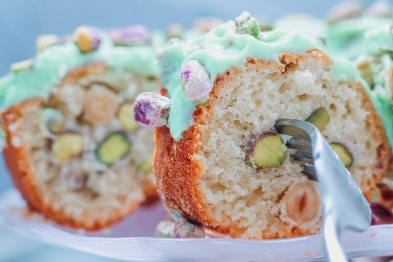 A slice of bundt cake with visible pistachios and hazelnuts, topped with green icing and whole pistachios, being cut with a fork.