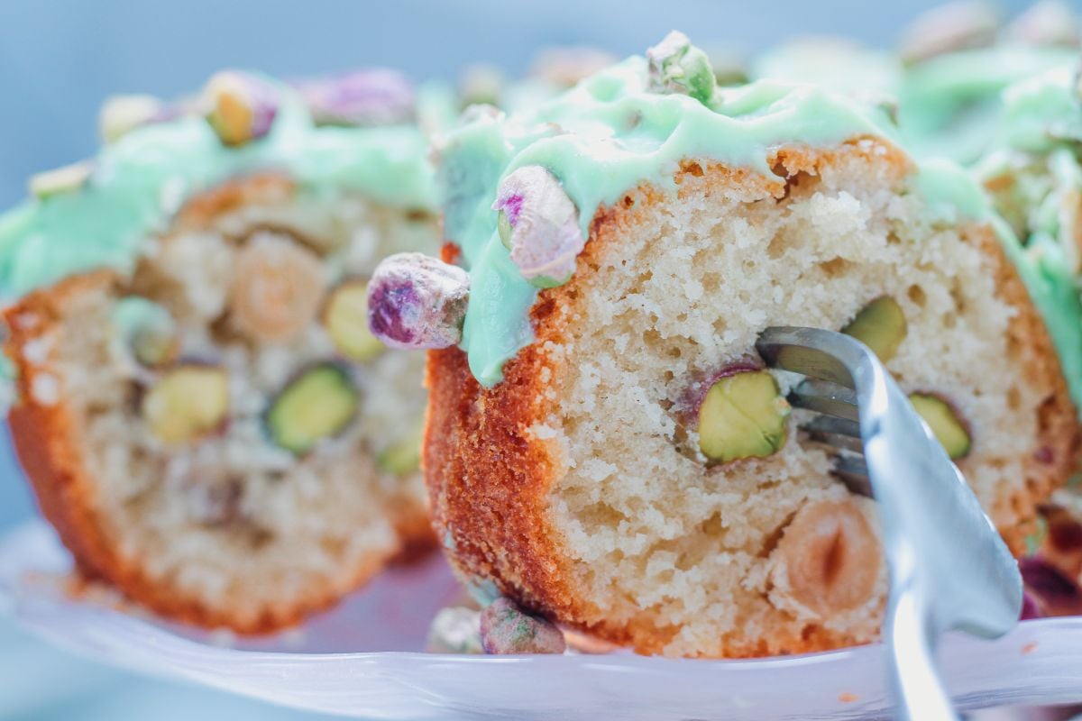 A close-up of a pistachio cake with green icing, topped and filled with whole pistachios, with a fork cutting a slice.