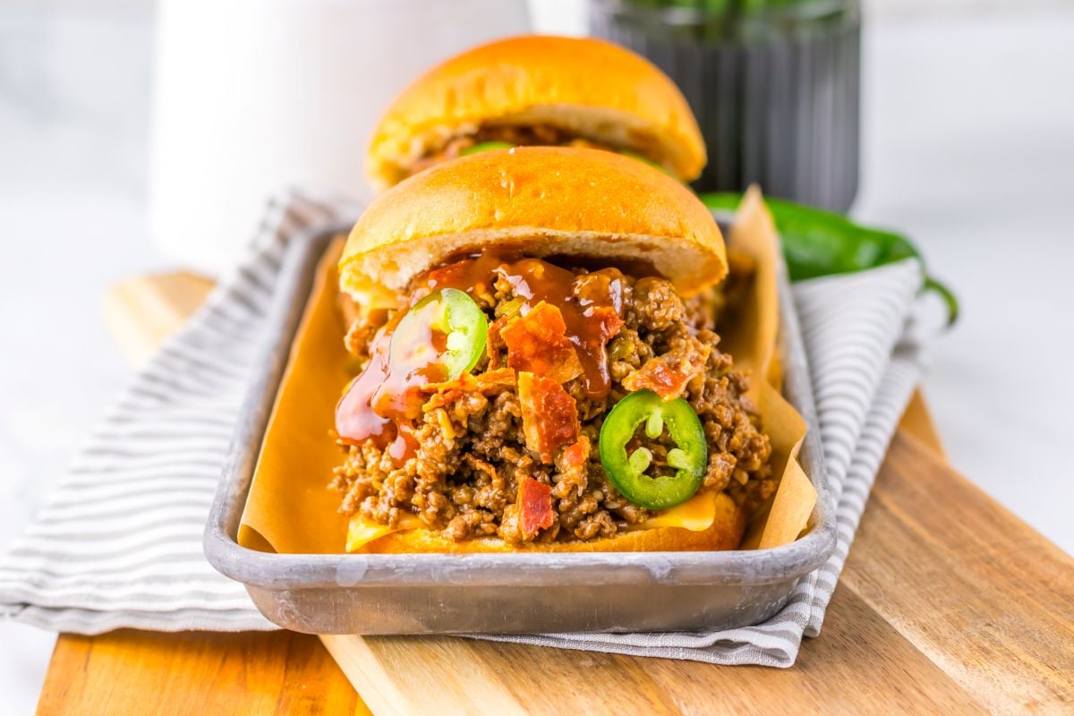 Two sloppy joe sandwiches topped with sliced jalapeños and bacon are served on a tray, placed on a striped napkin over a wooden board.